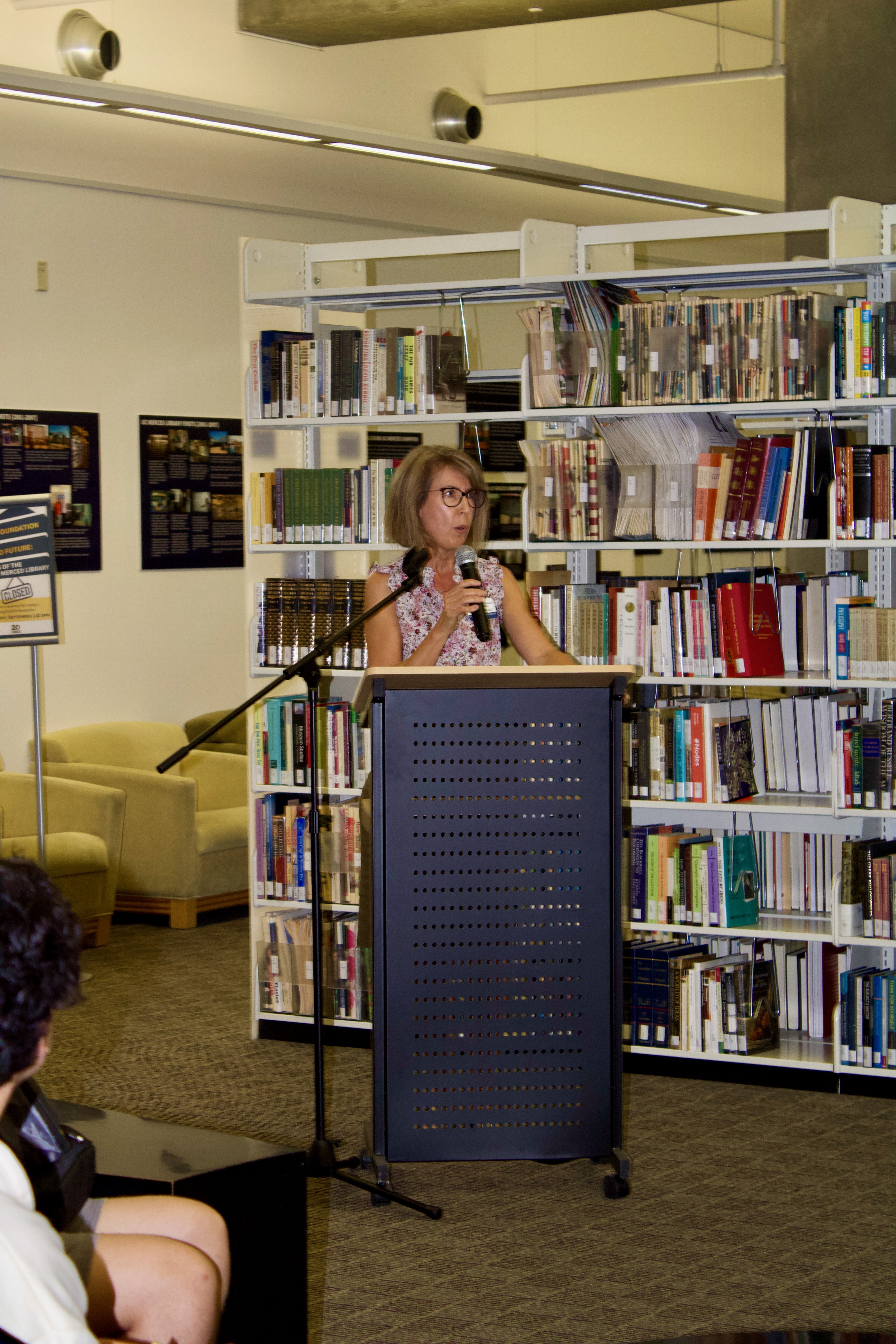 Individual stands behind podium and in front of bookstacks giving remarks