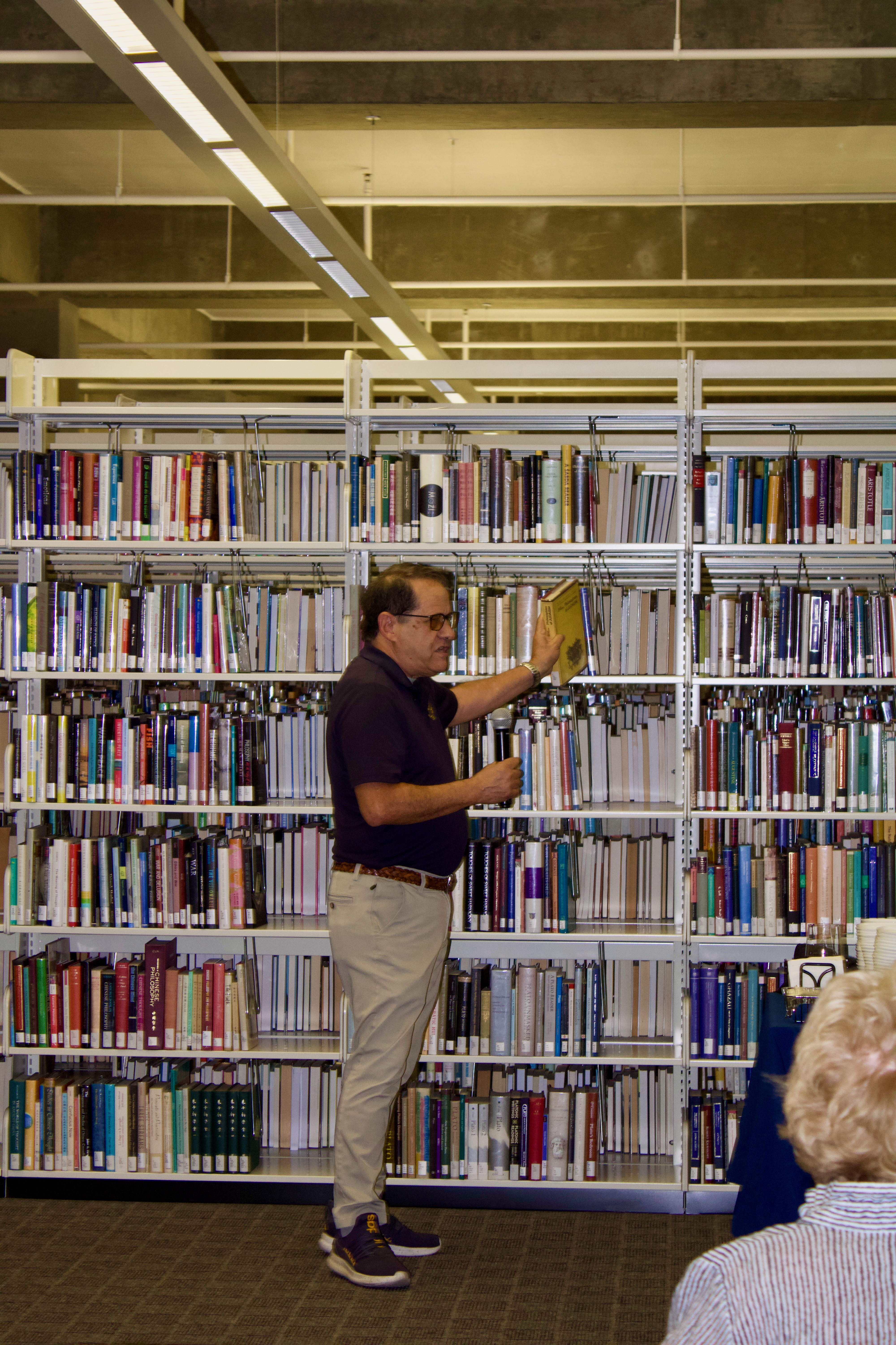 Side view of individual in front of book stacks, pulling a book from the shelf and speaking into a microphone