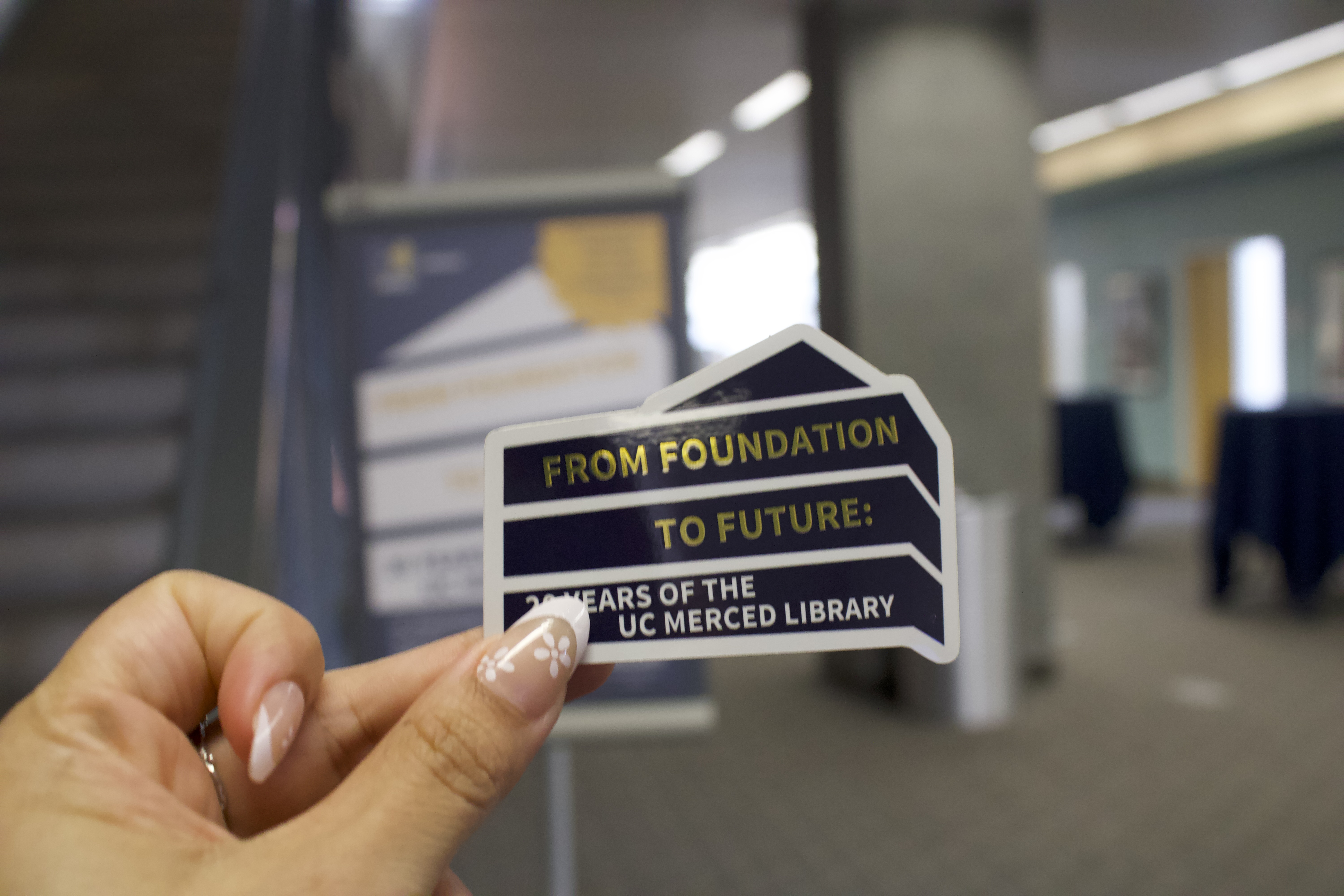Hand holding commerative exhibit sticker. From Foundation to Future: 20 Years of the UC Merced Library