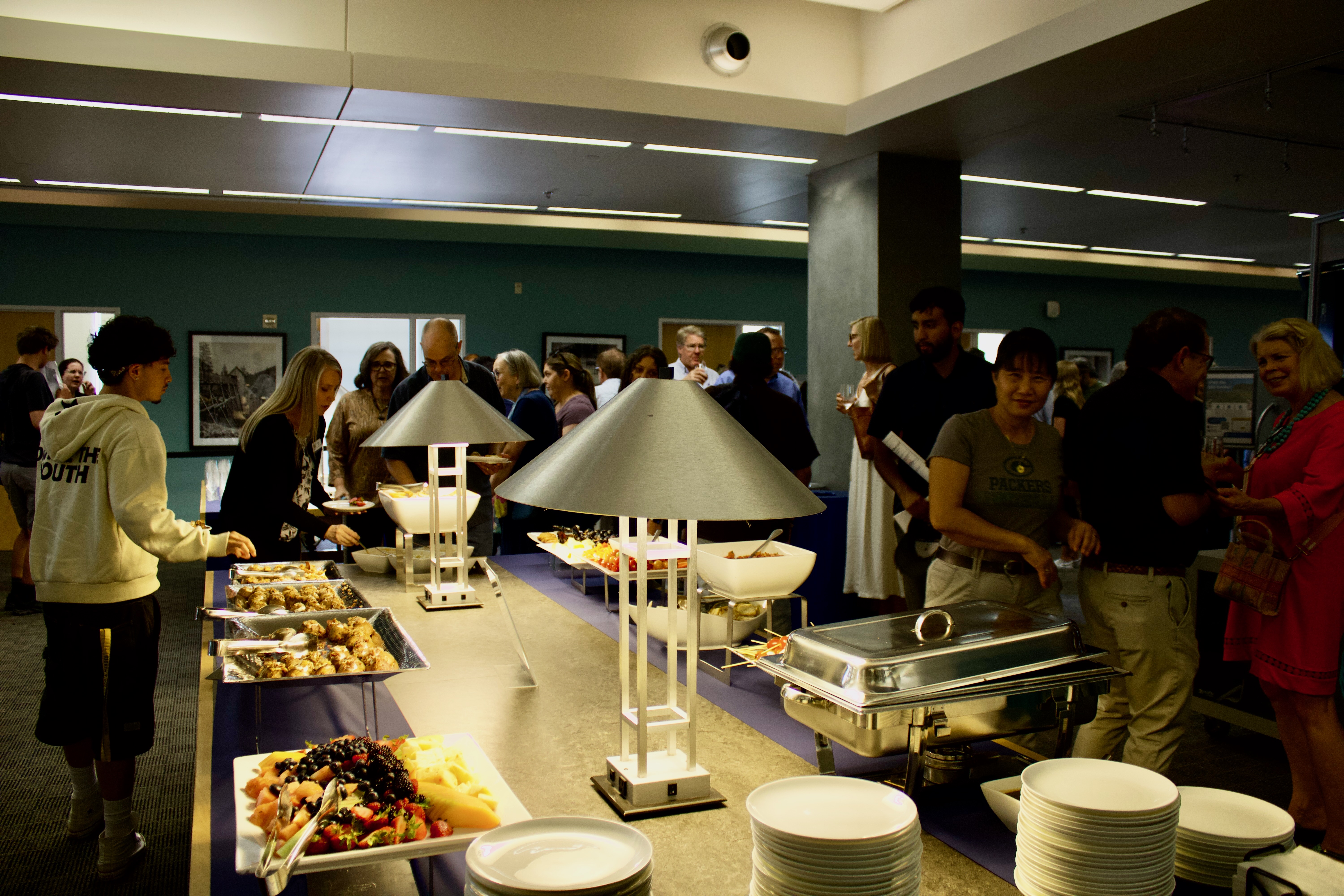 Food and plate set out on large table with lamps; individuals stand in background and eat