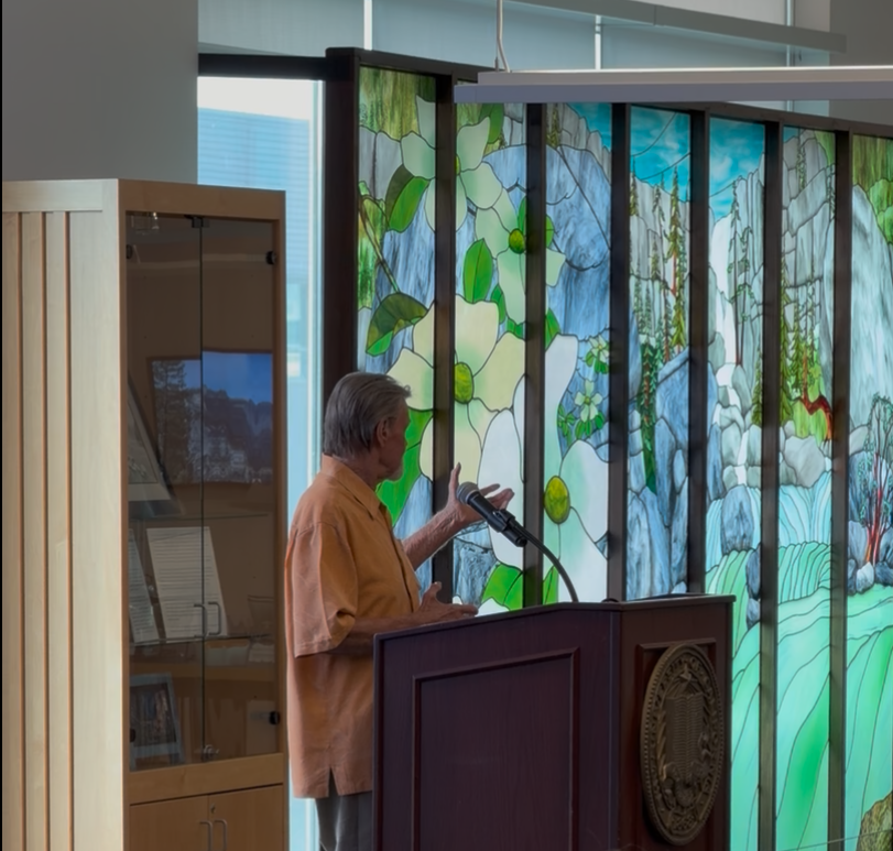 Bill Poulson speaks at podium in front of bookcase and spring Yosemite mural
