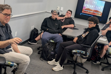 Mark Arax seated at left speaks to class with five students at right seated around a table.