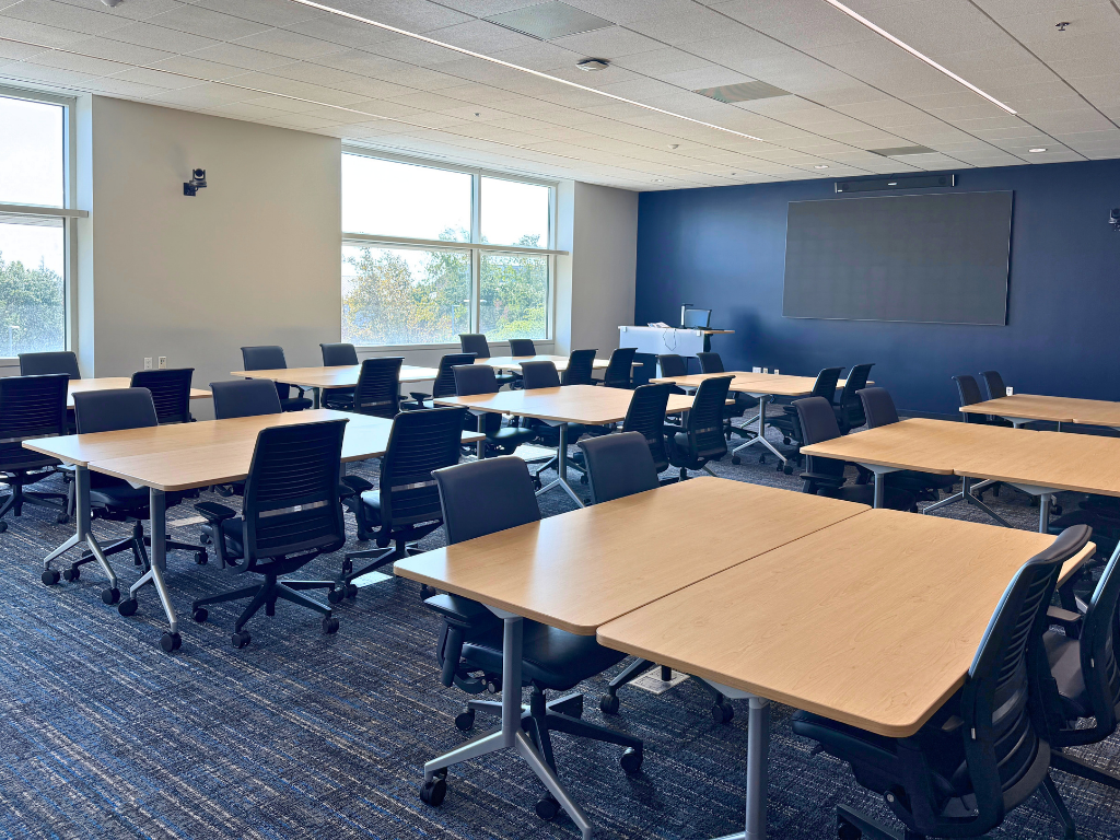 KL 232 room renovated with light-colored tables in pods of two with two dark chairs on each side; screen and podium at front of room