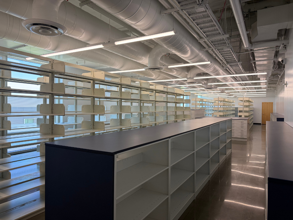 Expanded archives storage space with tall shelving at left and lower shelving in foreground with ducting overhead