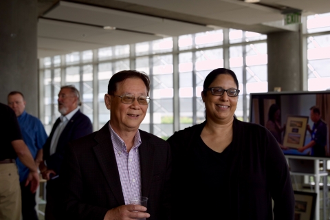 Two individuals stand and smile, guests in background at left, screen with slideshow at right