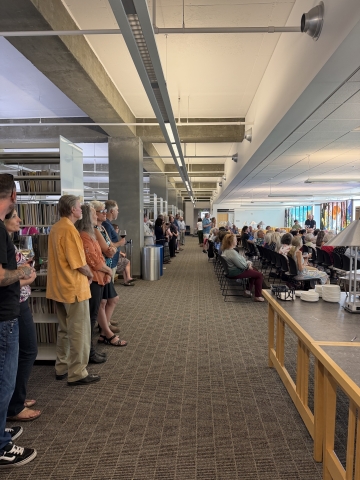 Individuals stand by book stacks and others are seated, facing a speaker
