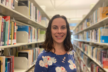 Image of Bronwen Maxson with short brown curly hair and a blue florap print top staning in front of a row of books on shelves