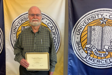 Photograph of Jim Dooley holding a certificate of recognition for his 20 year career service award, standing in front of a yellow and blue UC flag with the University of California seal