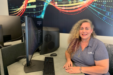 Woman with long curly hair sitting with arms folded on a desk with a computer on the desk