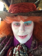 Woman dressed in orange wig, white face makeup and blue eyeshadow wearing a top hat 